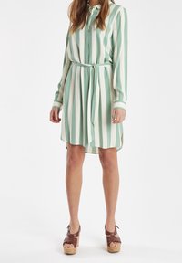 Abito camicia a righe verdi e bianche con maniche lunghe, chiusura a bottoni frontale, vita legata e design a pieghe. Accentuato con polsini a contrasto.
