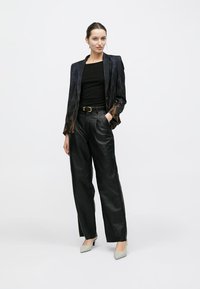 Blazer noir scintillant avec motifs dorés, haut noir ajusté, pantalon en cuir noir taille haute et talons gris clair.