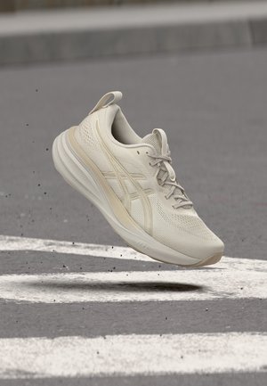 Chaussure de sport beige avec tige en mesh et semelle amortie en l'air au-dessus d'une route en asphalte avec des marquages blancs, montrant le mouvement et un design léger.