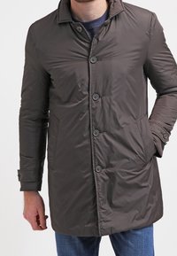 Tmavě hnědý, lehký, prošívaný kabát s límcem, šesti knoflíky a dvěma bočními kapsami. Má hladkou, lesklou texturu.