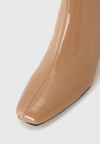 Bota de tacón alto y punta afilada en cuero patente nude brillante con detalle de costura vertical, textura lisa y suela negra.