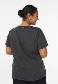 Frau mit dunklen Haaren zu einem Dutt gebunden, trägt ein dunkelgraues T-Shirt mit kurzen Ärmeln, von hinten vor einem schlichten weißen Hintergrund gesehen.