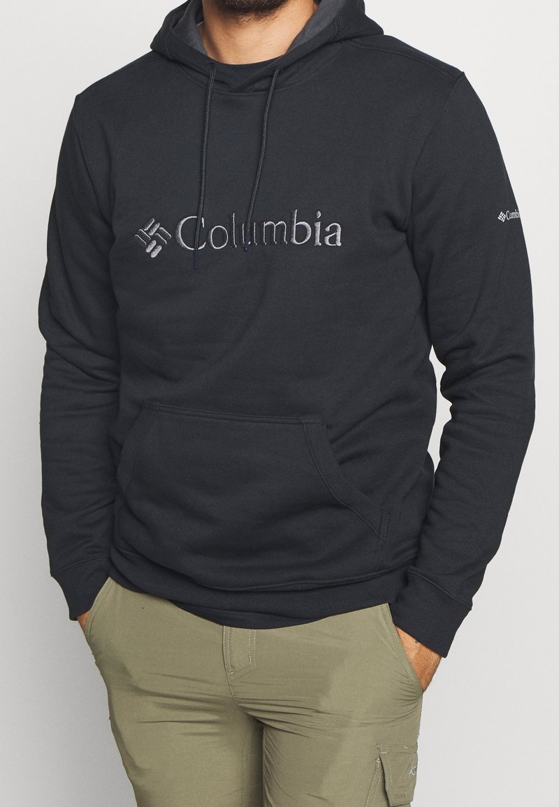 Homme portant un sweat à capuche noir Columbia avec poche avant et pantalon kaki, mains dans les poches, sur fond clair uni.
