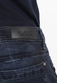 Bakifrån sedd person som bär mörkblå jeans med etiketten "BLEND Global Jeanskaker" i svart läder på midjebandet.