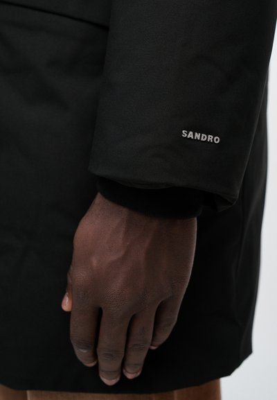 Crna izolirana jakna s rukavima koji imaju rebrasti rub i metalni logo "SANDRO". Tkanina izgleda glatko i blago sjajno.