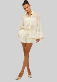 Vrouw staand met gekruiste benen, gekleed in een crèmekleurig, gestructureerd overhemd met lange mouwen en bijpassende korte broek, gecombineerd met enkellaarzen met bandje en bloemrijke oorbellen.