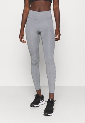Leggings gris avec une texture lisse, taille haute, et un détail de bande noire sur le côté. Associés à des chaussures de sport noires.