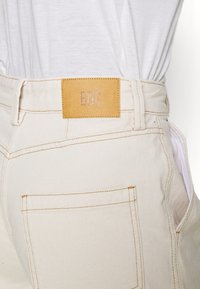 Beige denimbyxor har en tan läderetikett med "BDG" präglad, kontrasterande gul sömnad och detaljer på bakfickan.