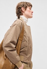 Rjavo bomber jakno z rebrastimi manšetami, stranskimi žepi in gladko teksturo, kombinirano s rjavim usnjenim ramo torbico. Pod njo bela visokokrožna majica.