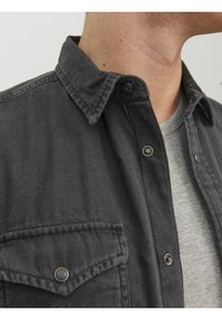 Camisa de botones gris oscuro con cuello, que cuenta con un bolsillo en el pecho y botones a presión plateados. Llevada sobre una camiseta de textura gris clara.