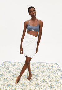 Grijze sportbeha, witte handdoek omgeslagen, staand op een bloemige deken met blauwe en gouden accenten. Glad texturen en neutrale kleuren.