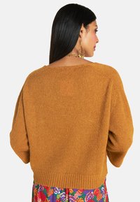 Senfgelbes gestricktes Sweatshirt mit einem cropped Design, lockeren Ärmeln und einem gerippten Saum. Getragen über einem floral gemusterten Kleid.