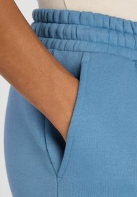 Light blue sweatpants with an elastic waistband, featuring a side pocket and soft, smooth texture. Close-up of hand resting in pocket.