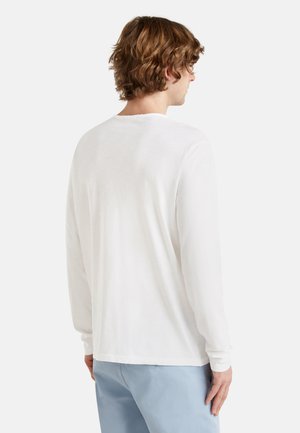 Homme aux cheveux bouclés de longueur moyenne portant une chemise blanche unie à manches longues et un pantalon bleu clair, tourné le dos contre un fond uni.