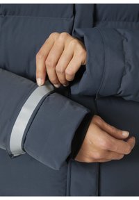 Námorní puffer bunda s hustou prošívanou strukturou; nastavitelné manžety s reflexním stříbrným pruhem; detailní záběr rukou upravujících rukáv.