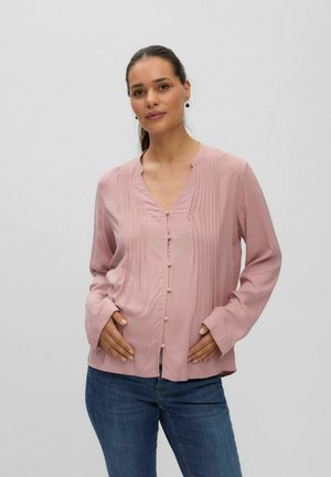 Femme aux cheveux foncés en queue de cheval basse porte un chemisier rose à manches longues avec des boutons et un jean bleu, debout sur un fond uni.