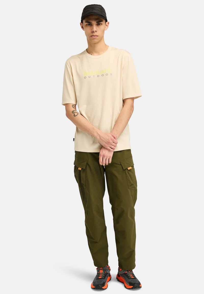 Beige T-Shirt mit kurzen Ärmeln und dem Aufdruck "Timberland Outdoor" in Grau und Grün. Olivgrüne Cargo-Hosen mit Taschen. Schwarze Kappe und Schuhe mit orangefarbenen Akzenten.
