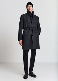 Cappotto lungo di lana grigio scuro con vita con cintura, collo alto e chiusura con bottoni. Abbinato a pantaloni neri aderenti e scarpe nere eleganti.