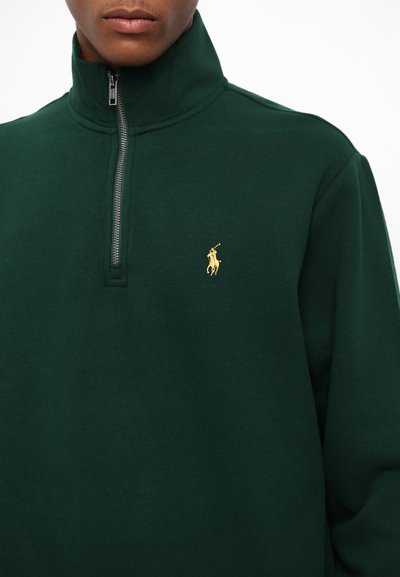 Tumši zaļa vilnas ceturtdaļas rāvējslēdzēja pulovers ar zelta izšuvumu polo logo kreisajā krūtī. Mīksta tekstūra, brīvs piegriezums. Rāvējslēdzēja detaļas ap kaklu.