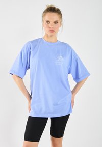 Lichtblauw oversized T-shirt van katoen, met korte mouwen en een ronde hals. Heeft een golfgrafiek en de tekst "AVONTUUR IS AAN HET BELLEN."