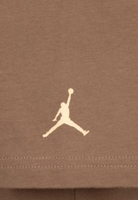 Hnědý bavlněný tričko s bílým logem "Jumpman", zobrazením postavy ve vzduchu, umístěným v dolním levém rohu. Viditelné švy.