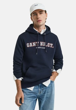 Navy hooded sweatshirt with front kangaroo pocket, white drawstrings, and contrast logo text. Worn over a white button-up shirt and blue jeans.