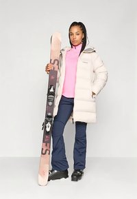 Kobieta w beżowej kurtce puchowej, różowej bluzce, granatowych spodniach i butach narciarskich trzymająca parę różowo-czarnych nart stojąca na jednolitym tle.