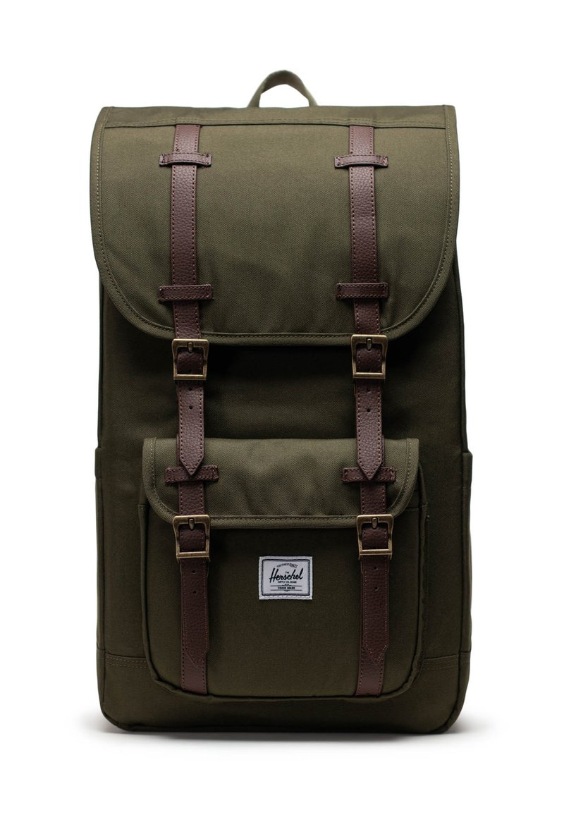 Zaino in tela verde oliva con patta, spallacci in pelle marrone, tasca frontale e un'etichetta con logo. Presenta una maniglia superiore per un facile trasporto.