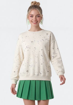 Smilende kvinde med blond hår i en topknold bærer en kremfarvet sweatshirt med pletter og et grønt plisseret skørt, accessoriseret med store grønne øreringe.