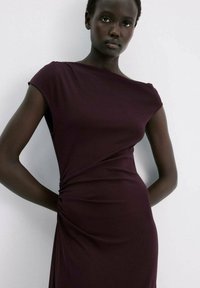 Vrouw met een donkere huid die een nauwsluitende, mouwloze paarse jurk draagt, staand tegen een effen lichte muur.