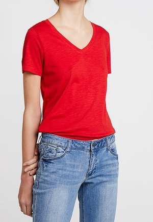 Osoba ubrana w prostą czerwoną koszulkę z krótkim rękawem i dekoltem w serek, włożoną do jasnoniebieskich jeansów, stojąca z jedną ręką za plecami.