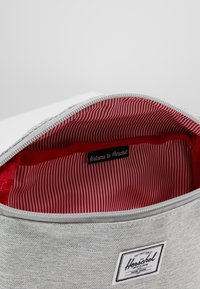 Grå tygpåse med inredning i röd och vit rand. Har en svart etikett med texten "Välkommen till Herschel" och ett vitt logopatch på utsidan.