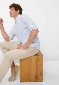 Lichtblauwe en witte gestreepte button-up shirt, beige slim-fit broek en tan instappers. Het model zit op een houten blok.