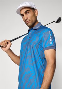 Homme portant une casquette blanche et une chemise de golf à motifs bleus, tenant un club de golf sur son épaule, regardant l'objectif avec une expression neutre.