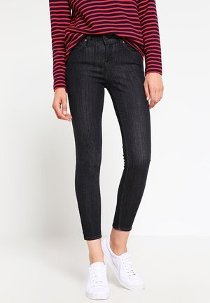 Persoon die donkere skinny jeans, een rood en marineblauw gestreept topje met lange mouwen en witte sneakers draagt, staand tegen een witte achtergrond.