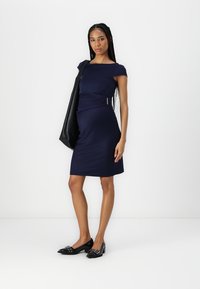 Vestido azul marinho até ao joelho, com mangas curtas, detalhe franzido na lateral e um leve accentuado metálico. Combinado com sapatilhas pretas e uma bolsa de tote.