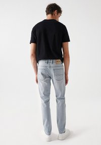 Camiseta negra de hombre, jeans azul claro con un lavado desgastado y zapatillas blancas. Los jeans tienen una etiqueta de cuero marrón en la parte trasera de la cintura.