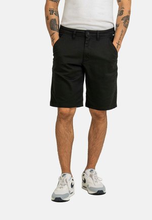 Man wearing black knee-length shorts, white t-shirt, and white Nike sneakers with black swoosh, standing with hands in pockets.