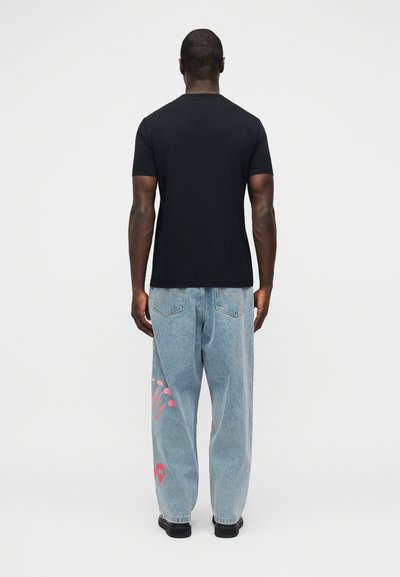 Άνδρας που στέκεται με την πλάτη προς τα εμπρός, φοράει απλό μαύρο κοντομάνικο πουκάμισο, ανοιχτό μπλε τζιν με ροζ γραφικά στο αριστερό πόδι και μαύρα παπούτσια.