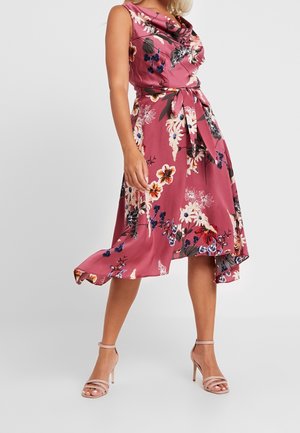 Femme portant une robe midi rose fleurie sans manches avec une ceinture à la taille et des sandales à talons haut couleur nude, debout sur un fond uni.