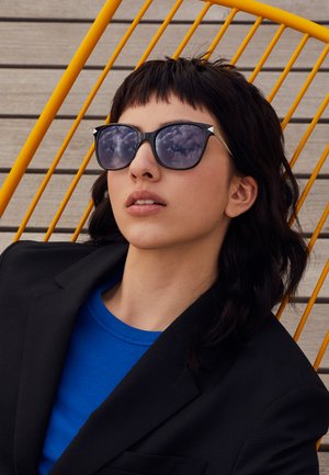 Black angular sunglasses with reflective tinted lenses, worn by a person in a black blazer and blue shirt, against a yellow chair.