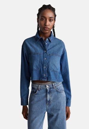 Chaqueta de mezclilla recortada en azul medio con mangas largas, cierre de botones, dos bolsillos en el pecho y detalles de costura visibles. Combinada con jeans de tiro alto.
