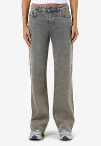 Sieviete valkā izbalējušas pelēkas taisnu kāju džinsas, baltu īsu topu un sudraba krāsas sporta apavus, stāvot ar rokām maigā pozīcijā sānos.