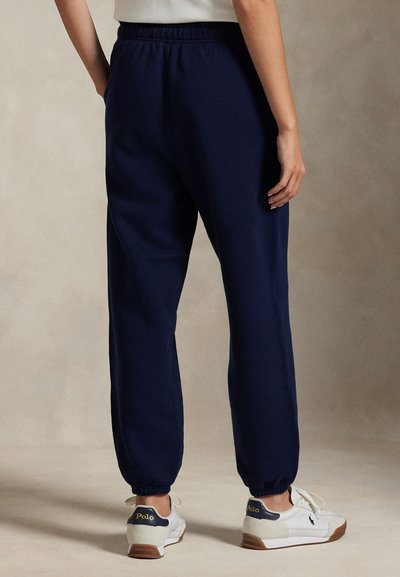 Άτομο που φοράει σκούρο μπλε παντελόνι jogger με ελαστική μέση και λευκά αθλητικά παπούτσια Polo, εμφανιζόμενο από πίσω πάνω σε ουδέτερο φόντο.