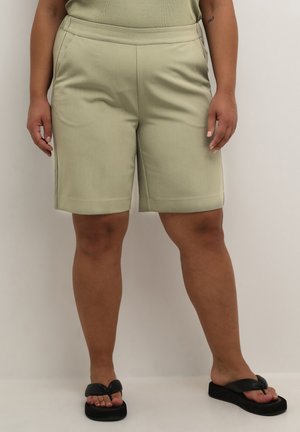 Pantaloni corti verde chiaro su misura con una texture liscia, tasche laterali e un orlo poco sopra il ginocchio. Indossati con infradito nere.