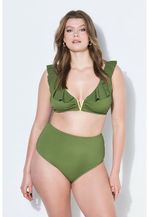 Frau, die ein olivgrünes Bikinioberteil mit Rüschen und hochgeschnittene Bikinihose trägt, steht vor einem weißen Hintergrund mit neutralem Gesichtsausdruck.