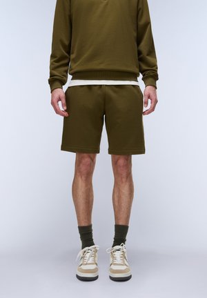 Hombre con sudadera verde oliva y pantalones cortos a juego, con calcetines oliva y zapatillas beige, de pie sobre un fondo neutro.