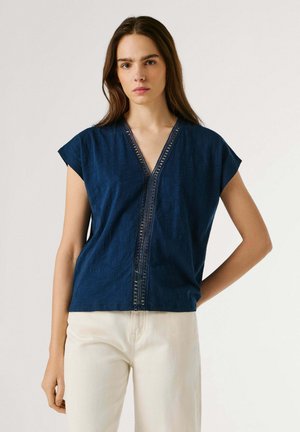 Donna che indossa un top senza maniche blu navy a scollo a V con rifiniture in pizzo, abbinato a pantaloni a vita alta color avorio, su sfondo semplice.