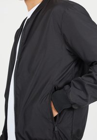 Svart bomberjacka i lätt material, med dragkedja fram, ribbade muddåkanter och sidofickor. Slät textur och figurnära design.