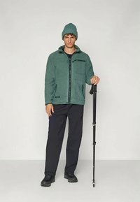 Fleecejakke i grønn med glidelås foran, to lommer og svarte detaljer; brukt sammen med marineblå bukser. Svart turstav holdt i høyre hånd.